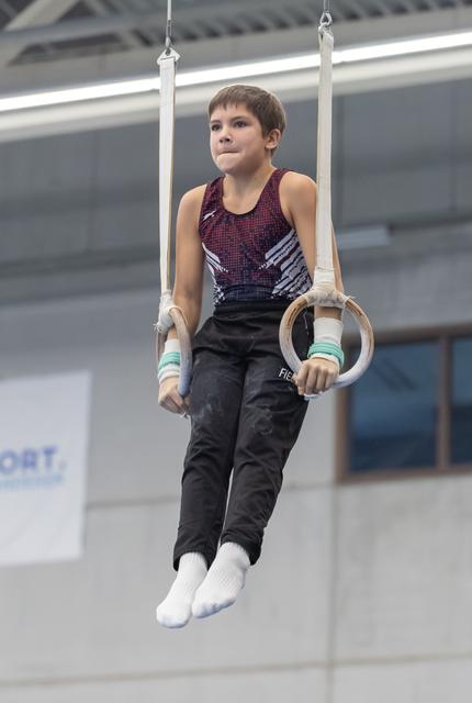 Young male gymnast holds position on rings apparatus, wearing burgundy and black leotard with focused expression