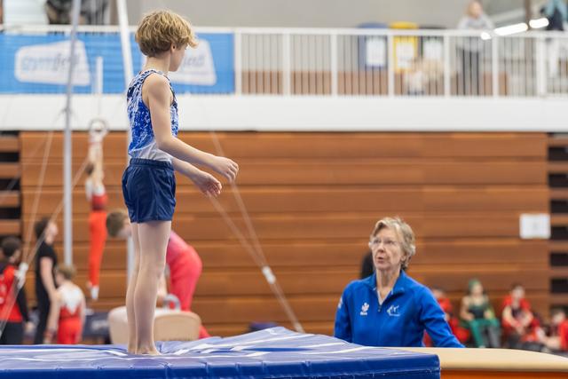 Young gymnast stands on vault apparatus while female coach in blue jacket observes from below during training session