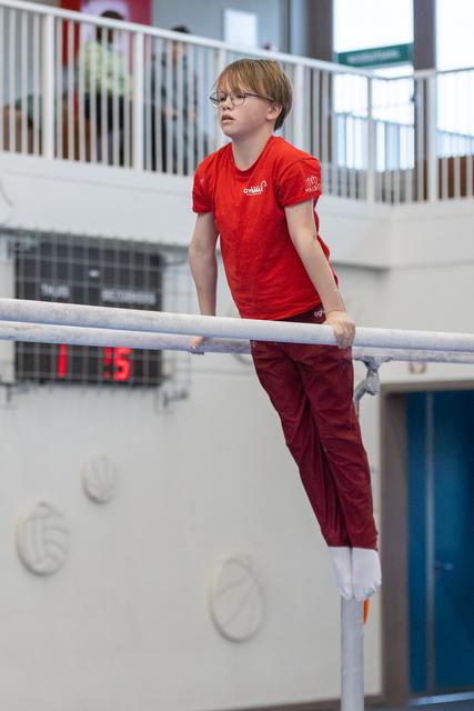 Young gymnast in red shirt and glasses performs on parallel bars, displaying focus and determination in training facility