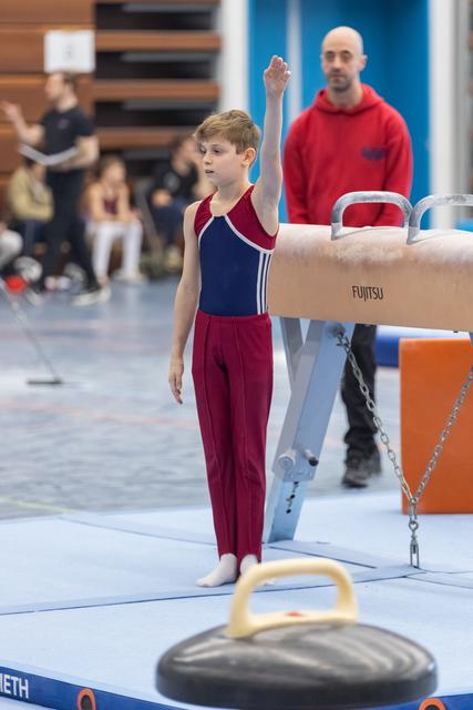 Young gymnast stands beside pommel horse with raised hand, focused expression, while coach observes in background