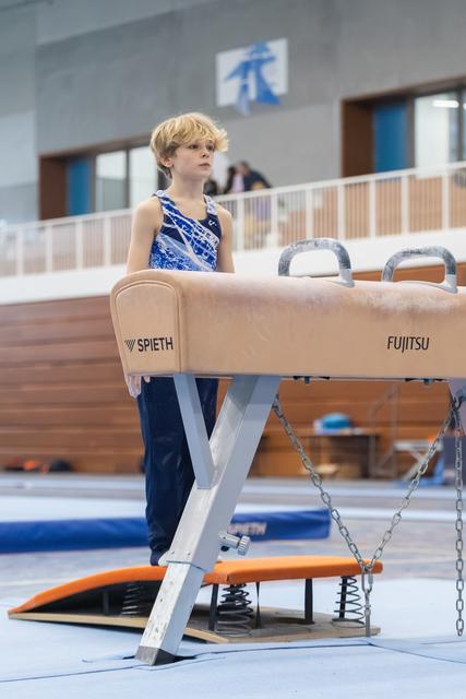 Young gymnast with blonde hair stands beside pommel horse, gazing upward with focused expression in training facility