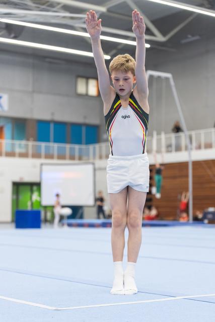 Young red-haired gymnast in white and rainbow-trimmed leotard stands with arms raised overhead in starting position