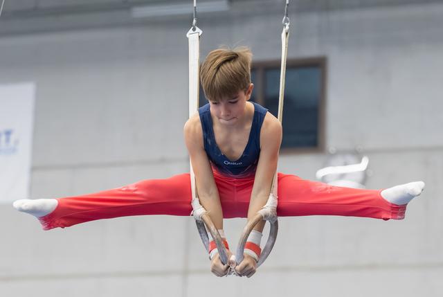 Young gymnast performs horizontal split on still rings, demonstrating strength and flexibility in red pants and blue leotard