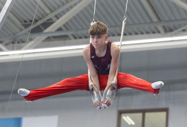 Male gymnast performs straddle position on still rings, displaying strength and control in indoor training facility