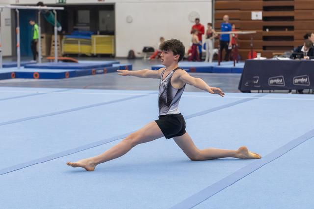 Young gymnast executes a graceful split leap during floor routine, arms extended, in front of spectators and judges