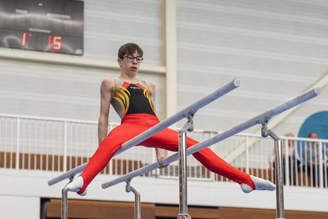 Young gymnast executes a split position on parallel bars, wearing red pants and black-orange top, focused expression