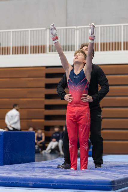 Young gymnast in red ombre outfit raises arms in triumph while coach stands supportively behind on blue mat in gymnasium