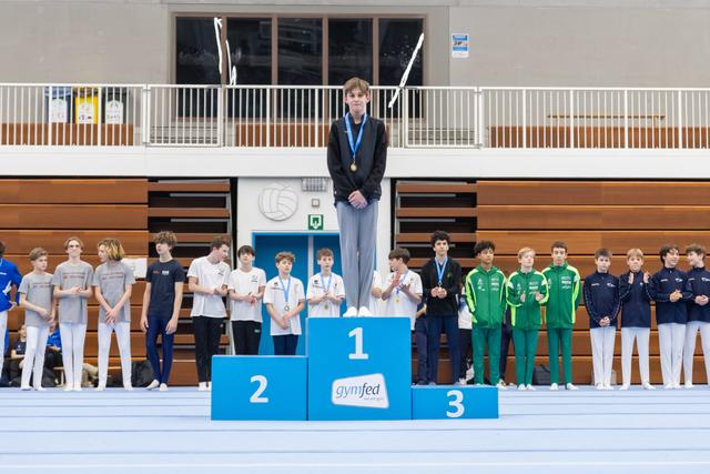 Young gymnast standing on first place podium wearing medal, with second and third place teams lined up beside podium