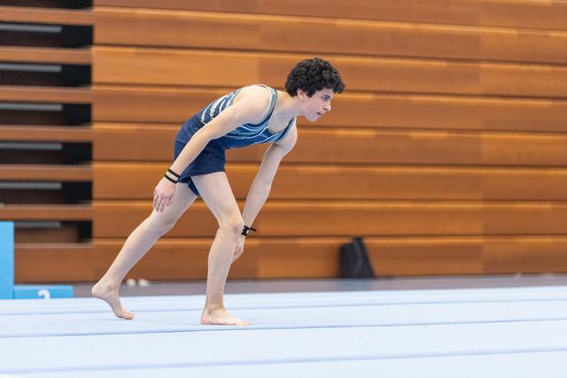Young male gymnast in blue leotard performs a deep lunge on floor mat, demonstrating flexibility and concentration