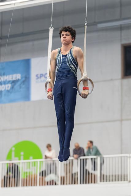 Male gymnast suspended on rings in focused concentration, wearing navy uniform with white hand guards during routine