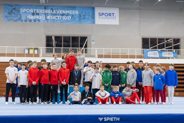 Large group of young male gymnasts in colorful training gear posing together on a blue mat in an indoor gymnasium