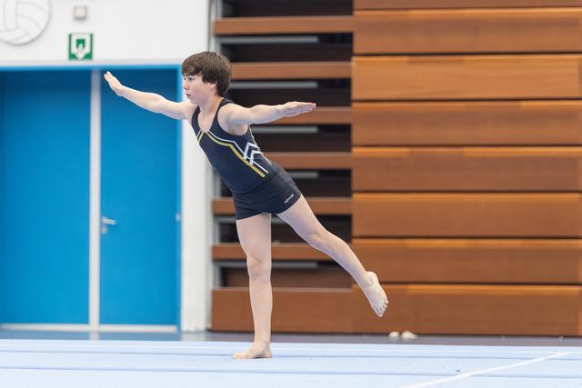 Young gymnast performing an arabesque during floor routine, arms extended gracefully in navy leotard at indoor training facility