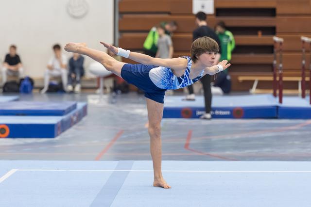 Young gymnast performs a T-pose balance on floor, arms extended horizontally, demonstrating flexibility and control