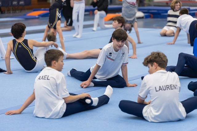 Young gymnasts in white polo shirts perform floor stretches during warmup, with names like 'AXEL' and 'LAURENT' on their backs