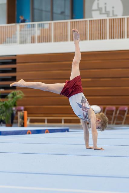 Young male gymnast performs a one-armed handstand with legs split on a blue gymnastics floor mat indoors.