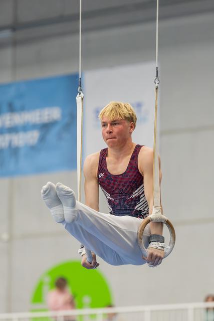 Young male gymnast grips still rings with intense concentration, legs tucked, competing in an indoor gymnastics hall.