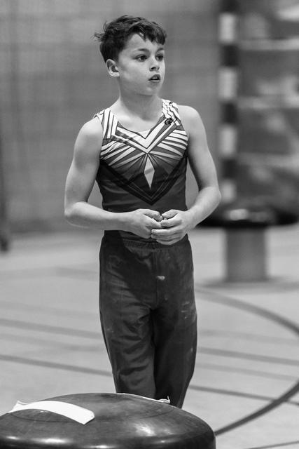 Young gymnast stands composed beside vault, hands clasped, wearing black and white striped leotard in training facility