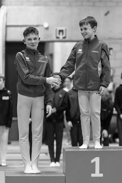 Two young gymnasts shake hands on podium, first and second place, both smiling in matching team jackets