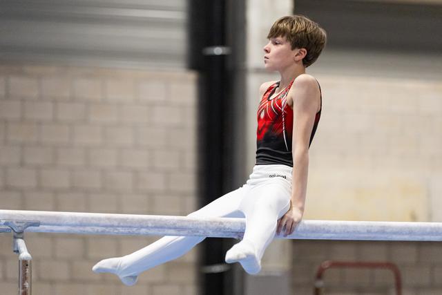 Young gymnast in red and black leotard sits on parallel bars with focused expression during routine