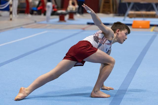Young gymnast performs a lunge position during floor routine, arms extended with focused expression on blue mat