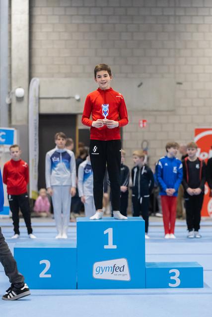 Young male gymnast in red jacket stands on first place podium holding medal, smiling proudly at gymnastics event