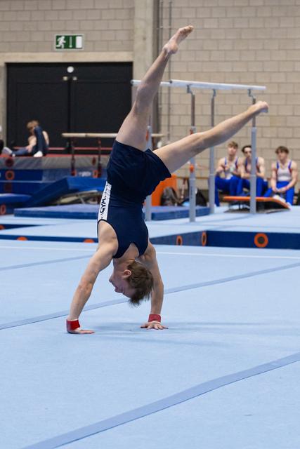 Male gymnast executing a one-armed handstand during floor routine, demonstrating strength and balance on blue mat