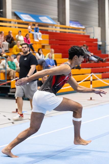 Male gymnast in red and white leotard performs dramatic lunge pose on floor mat, arms extended, with spectators in bleachers