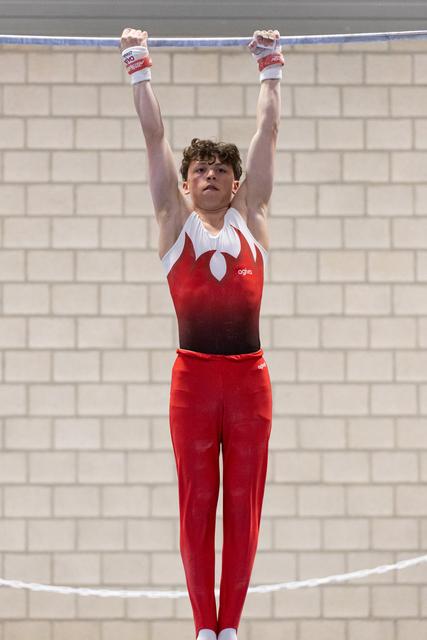 Young male gymnast hangs from horizontal bar in red and white leotard, arms extended overhead, focused expression