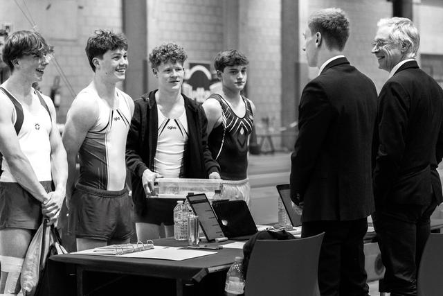 Four young gymnasts in competition attire stand smiling at a table, conversing with two officials in dark suits