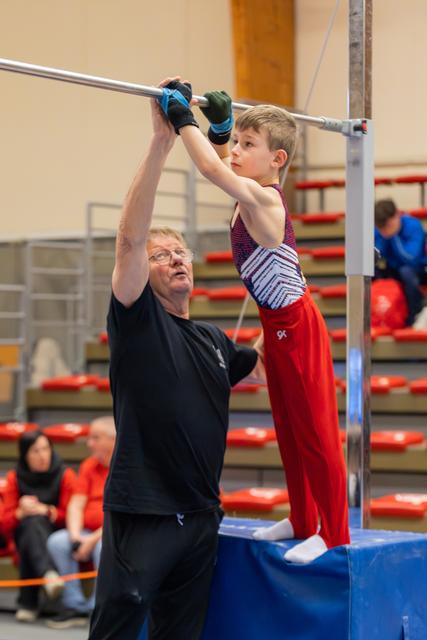 An older coach steadies a focused young boy gripping the high bar during a gymnastics competition in a red-seated arena.