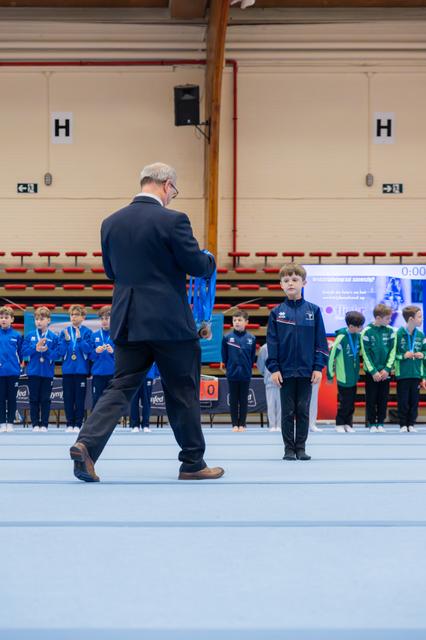 An official in a suit approaches a young boy in a blue tracksuit to present a medal during a gymnastics awards ceremony.