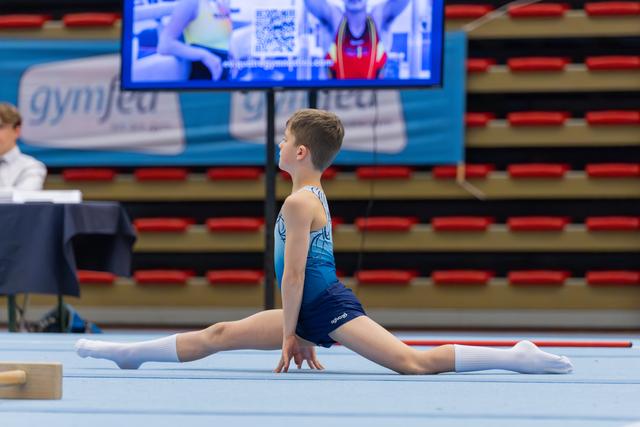 Young male gymnast performs a perfect split on the floor, back arched, focused expression during an indoor gymnastics meet.