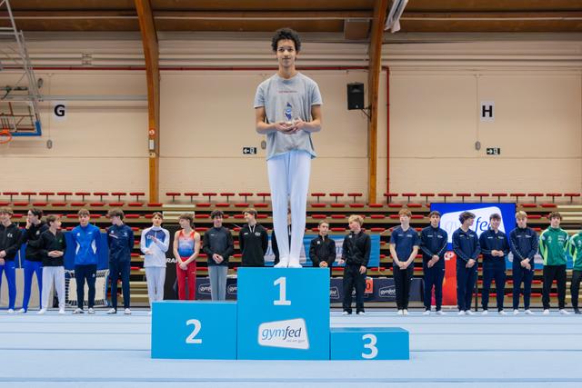 Young male gymnast stands proudly on the first-place podium, holding a trophy at a Gymfed indoor gymnastics event.