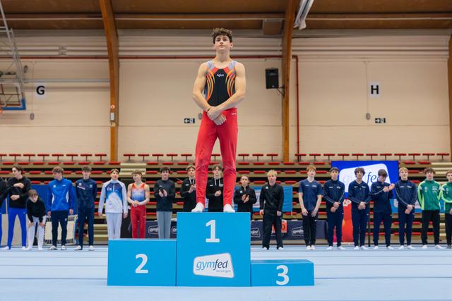 Young male gymnast stands proudly on the first-place podium at a gymfed indoor gymnastics championship, arms crossed.
