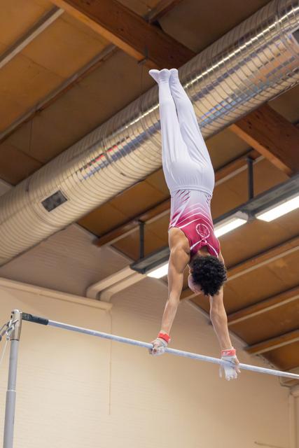 Male gymnast holds a perfect handstand on the high bar, body fully extended, in an indoor gymnasium.