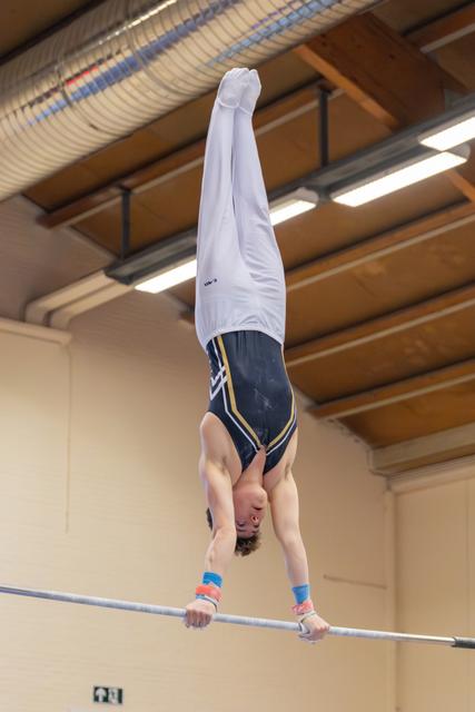 Young male gymnast performs a handstand on the horizontal bar, body fully extended upside down in an indoor gymnasium.