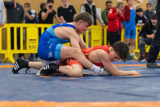 Young wrestler in blue singlet dominates opponent in red, pressing him down on the mat during an indoor match.