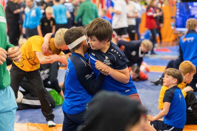 Two young boys in blue Jako jerseys lock grips, faces close, intense concentration during a busy indoor wrestling event.