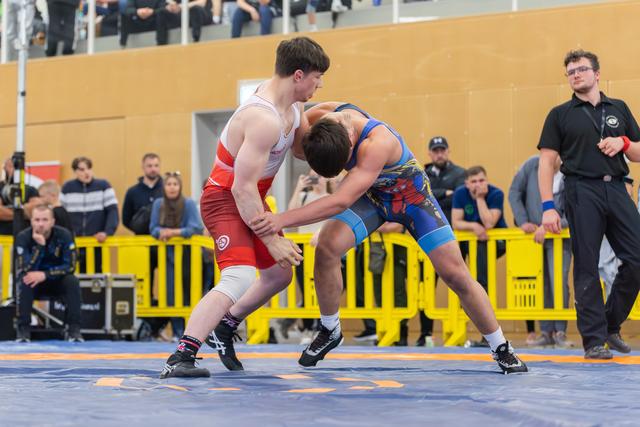 Two young wrestlers locked in intense grappling, one in red singlet, one in blue, battling on a blue competition mat.