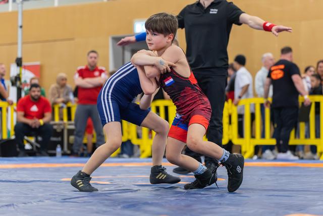 Two young wrestlers lock grips on the mat, focused and determined, as a referee signals in the background.