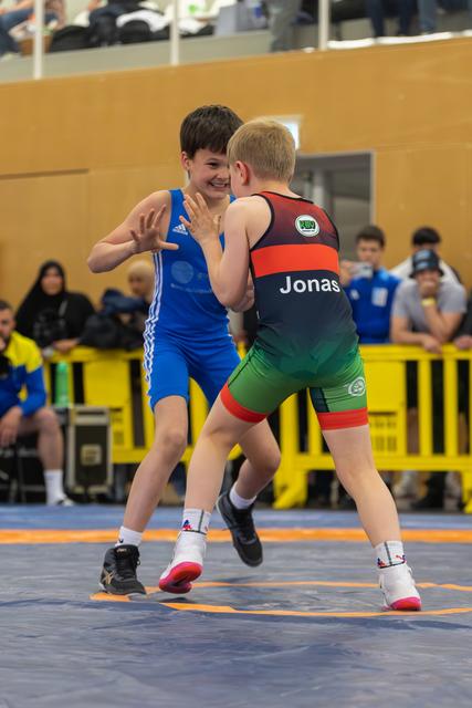 Smiling boy in blue Adidas singlet faces Jonas in black-red-green singlet on blue mat, indoor gym.