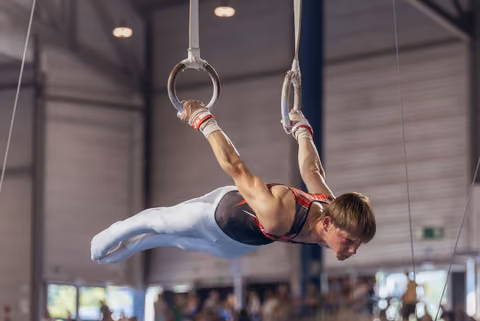 Young male gymnast holds an iron cross on still rings, body horizontal, intense concentration, indoor sports hall.