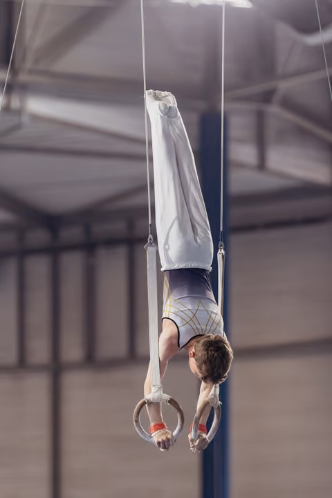 Male gymnast holds a straight body inversion on still rings, wearing white pants and a decorated leotard, in an indoor gym.