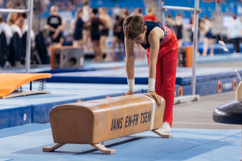 Young male gymnast leans intently over a pommel horse, hands on apparatus, focused before his routine in a busy gym.