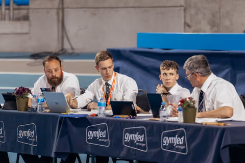 Four male judges in white shirts review scores on laptops at a Gymfed-branded table during a gymnastics competition.