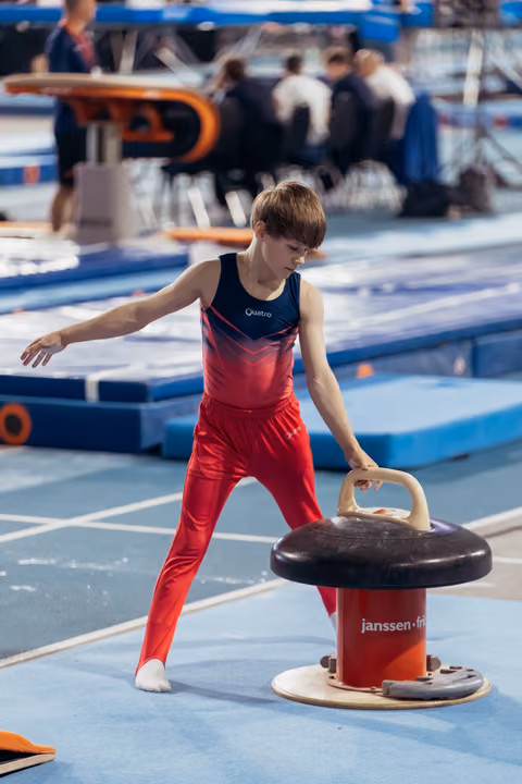 Focused young male gymnast in red uniform grips pommel horse handles, preparing for routine in blue gymnasium.