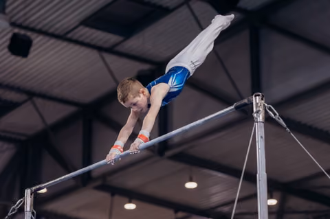 Young male gymnast executes a daring horizontal bar release, body fully extended, focused expression in an indoor gym.
