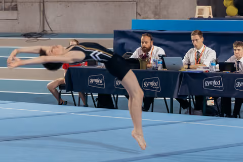 Young gymnast performs a powerful leap on the floor exercise mat while Gymfed judges observe attentively at their table.