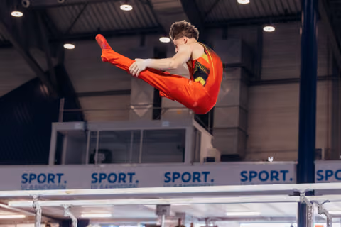 Male gymnast in red suit performs a dynamic straddle jump above parallel bars, legs fully extended, intense focus.