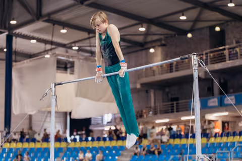 Young male gymnast grips the high bar with focused intensity, body extended, in a packed indoor gymnastics arena.